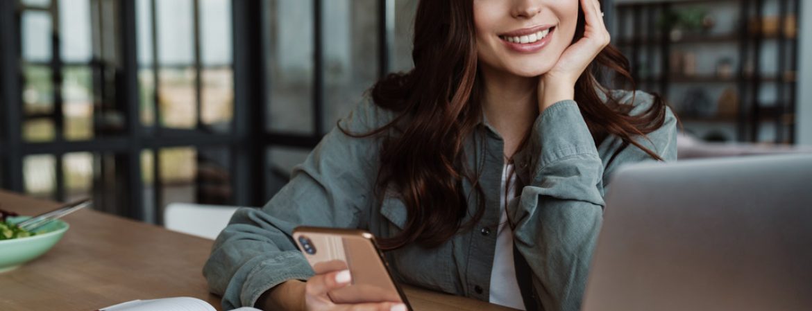 Beautiful smiling woman using cellphone while working with laptop Beautiful smiling woman using cellphone while working with laptop indoors
