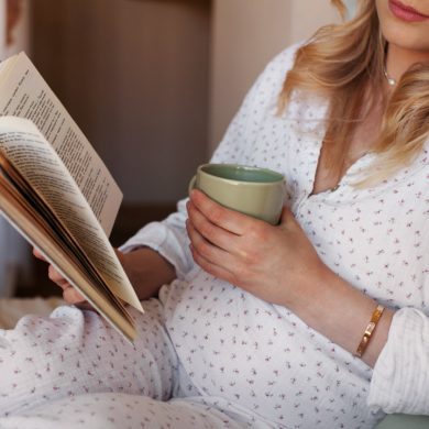 Close up Pregnant woman sitting at home in bed, reading book and drinking tea or water from cup